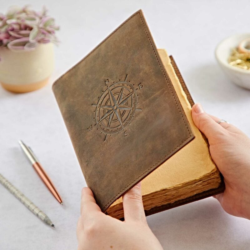 Hands holding an open, Personalised Compass Buffalo Leather Refillable Journal. A pen and plant are visible in the background.