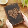 A person holds a Personalised Buffalo Leather Refillable Journal with an intricate heart design, marked with "E & J" at the bottom right, on a wooden table beside a green plant and a cup of coffee.
