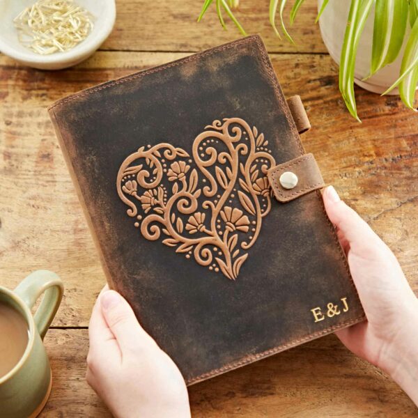A person holds a Personalised Buffalo Leather Refillable Journal with an intricate heart design, marked with "E & J" at the bottom right, on a wooden table beside a green plant and a cup of coffee.