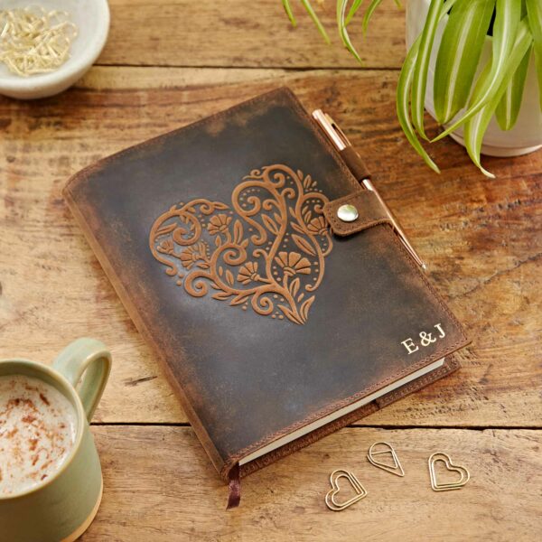A Personalised Heart Buffalo Leather Refillable Journal with an ornate heart design on the cover, a pen loop, gold initial letters "E & J," lies on a wooden table alongside a cup of coffee, gold paperclips, and a plant. This refillable journal combines elegance and practicality perfectly.