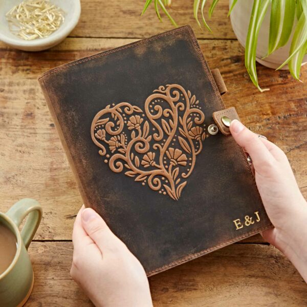 Hands holding a Personalised Heart Buffalo Leather Refillable Journal with an intricately embossed heart design and the initials "E & J" on a wooden table. A plant, a mug, and a bowl of paperclips are in the background.