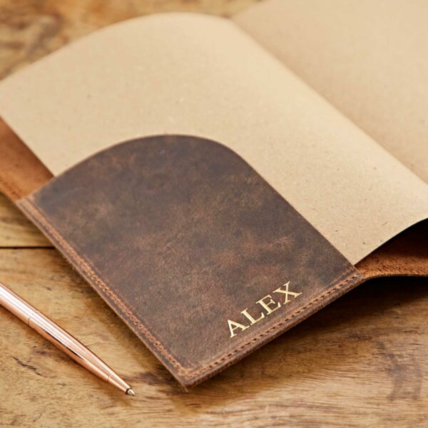 A Personalised Buffalo Leather Refillable Journal with the name "ALEX" embossed on the inside pocket lies open on a wooden table beside a rose gold pen.