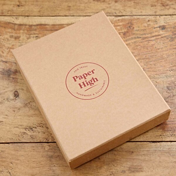 A brown cardboard box labeled "Paper High" sits on a wooden surface. The label reads "Fair Trade, Handmade & Sustainable" in a circular design, hinting at the Personalised Buffalo Leather Refillable Journal inside.