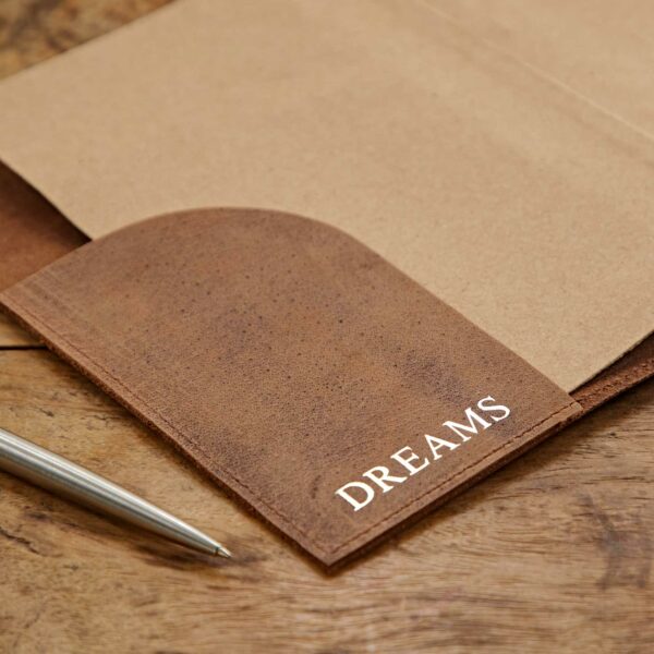 A brown Personalised Buffalo Leather Refillable Tree of Life Journal with the word "DREAMS" written on the cover sits on a wooden surface beside a silver pen.
