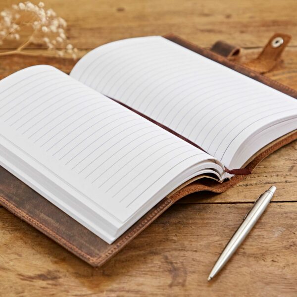 An open lined Personalised Buffalo Leather Refillable Tree of Life Journal lies on a wooden surface, accompanied by a silver pen.