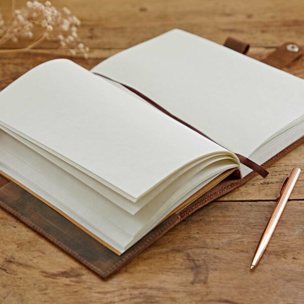A Personalised Heart Buffalo Leather Refillable Journal lies open on a wooden table, with blank pages exposed. A gold-colored pen is placed next to it, ready to be used in this refillable notebook.