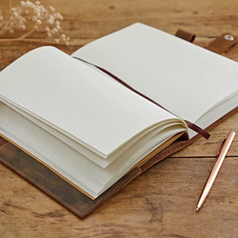 A Personalised Heart Buffalo Leather Refillable Journal lies open on a wooden table, with blank pages exposed. A gold-colored pen is placed next to it, ready to be used in this refillable notebook.
