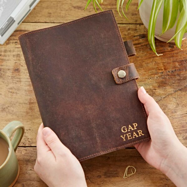 A person holding a Personalised Buffalo Leather Refillable Journal labeled "GAP YEAR" on a wooden table, with a green mug, a plant, and a small paperclip nearby.