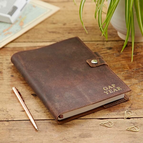 A Personalised Buffalo Leather Refillable Journal labeled "Gap Year" sits on a wooden table with a pen, paperclips, a potted plant, and a map.