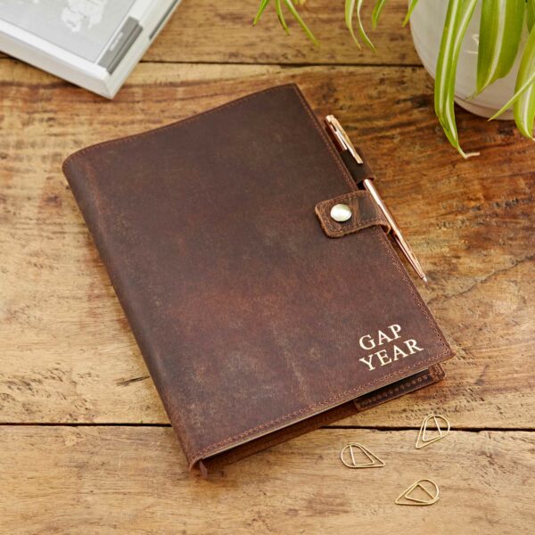 A brown Personalised Buffalo Leather Refillable Journal labeled "GAP YEAR" with a pen clipped to it rests on a wooden table next to some paper clips and a green plant. This personalised, refillable journal adds a touch of elegance to your gap year adventures.