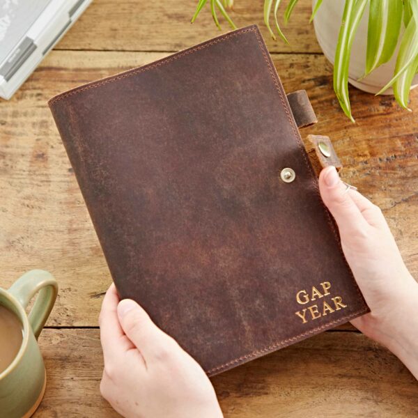 Person holding a Personalised Buffalo Leather Refillable Journal with the words "GAP YEAR" embossed on the cover, next to a green plant and a cup of coffee on a wooden surface.