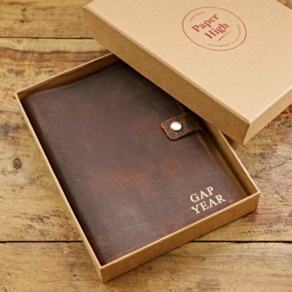 A sustainably sourced Personalised Buffalo Leather Refillable Journal labeled "GAP YEAR" rests in an open cardboard box on a wooden surface. The lid of the box reads "Paper High" and "Handmade in India," highlighting its personalised touch.