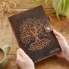 Hands holding a Personalised Buffalo Leather Refillable Tree of Life Journal with an embossed Tree of Life design and the word "FAITH" on the cover, placed on a wooden surface near a plant and a cup of coffee.