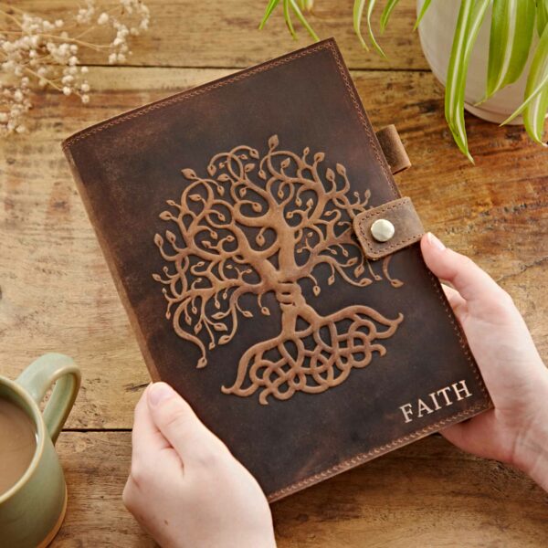 Hands holding a Personalised Buffalo Leather Refillable Tree of Life Journal with an embossed Tree of Life design and the word "FAITH" on the cover, placed on a wooden surface near a plant and a cup of coffee.