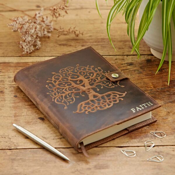 A Personalised Buffalo Leather Refillable Tree of Life Journal sits on a wooden table next to a pen, paper clips, and a green potted plant.