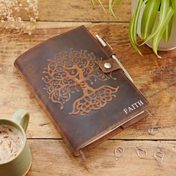 A Personalised Buffalo Leather Refillable Tree of Life Journal with the word "FAITH" on the cover rests on a wooden surface. Nearby, there is a green mug, some metal bookmarks, and a plant, completing the serene setting.
