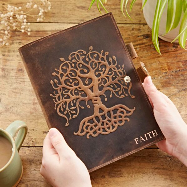A person holds a Personalised Buffalo Leather Refillable Tree of Life Journal engraved with an intricate Tree of Life pattern and the word "FAITH," placed near a green plant and a mug of coffee on a wooden table.