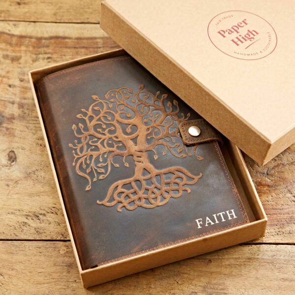 A Personalised Buffalo Leather Refillable Tree of Life Journal with an embossed Tree of Life design and the word "FAITH" lies partially in an open cardboard box on a wooden surface. The box lid is labeled "paper High Handmade & Fair Trade.