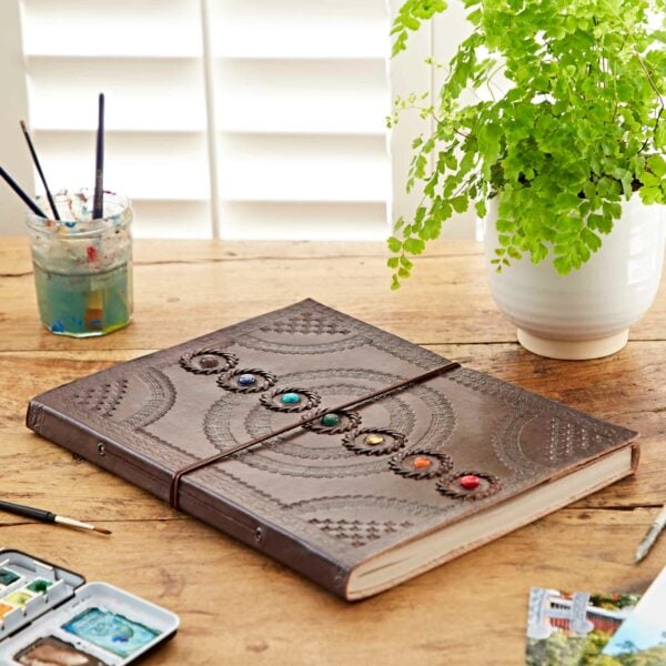 A large Indra Chakra Embossed Leather Album with Semi-Precious Stones lies on a wooden table next to a jar of paintbrushes, a watercolor palette, and a potted plant.
