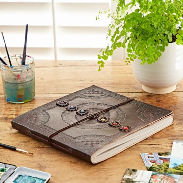 An Indra Chakra Embossed Leather Album with Semi-Precious Stones, intricately embossed with a circular Indra Chakra pattern and colored accents, rests on a wooden table near paintbrushes, a glass jar, and a potted fern.