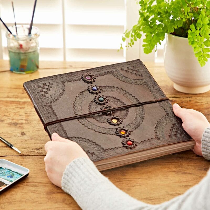 Person holding a large Indra Chakra Embossed Leather Album with Semi-Precious Stones on a wooden desk near a potted plant and watercolor painting supplies.