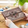 A person holds an intricately designed Indra Chakra Circle Embossed Leather Album with Semi-Precious Stones on a wooden table beside photos, scissors, and a plant.
