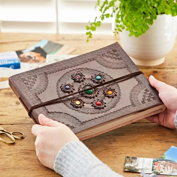A person holds an intricately designed Indra Chakra Circle Embossed Leather Album with Semi-Precious Stones on a wooden table beside photos, scissors, and a plant.