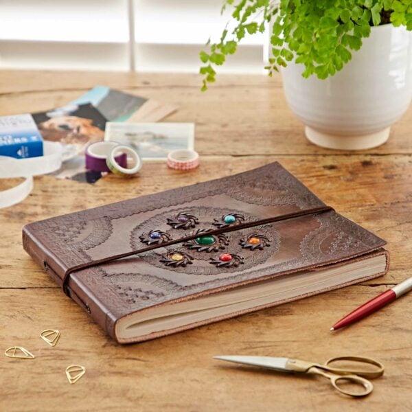 A closed Indra Chakra Circle Embossed Leather Album with Semi-Precious Stones sits on a wooden table, surrounded by washi tapes, scissors, a pen, a potted fern, photos, and decorative paper clips.