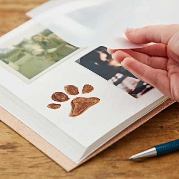 A hand lifts a sheet in the Indra Chakra Circle Embossed Leather Album with Semi-Precious Stones on a wooden table, revealing a page with a dog's paw print and two photos. A pen lies nearby.