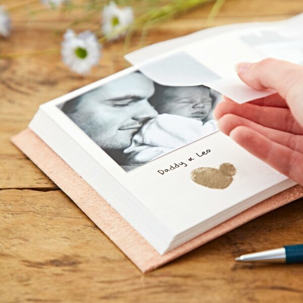 A hand is lifting a photo in an open Indra Chakra Circle Embossed Leather Album with Semi-Precious Stones labeled "Daddy & Leo" beside a pen and white flowers on a wooden table.