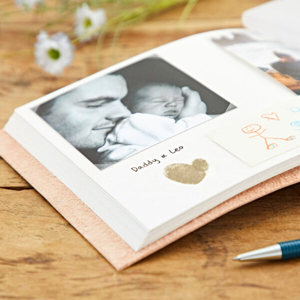 An open Indra Chakra Circle Embossed Leather Album with Semi-Precious Stones rests on a wooden table, featuring a black and white picture of a man and a baby labeled "Daddy & Leo." Additional drawings and a silver heart decoration are visible. A pen lies nearby.