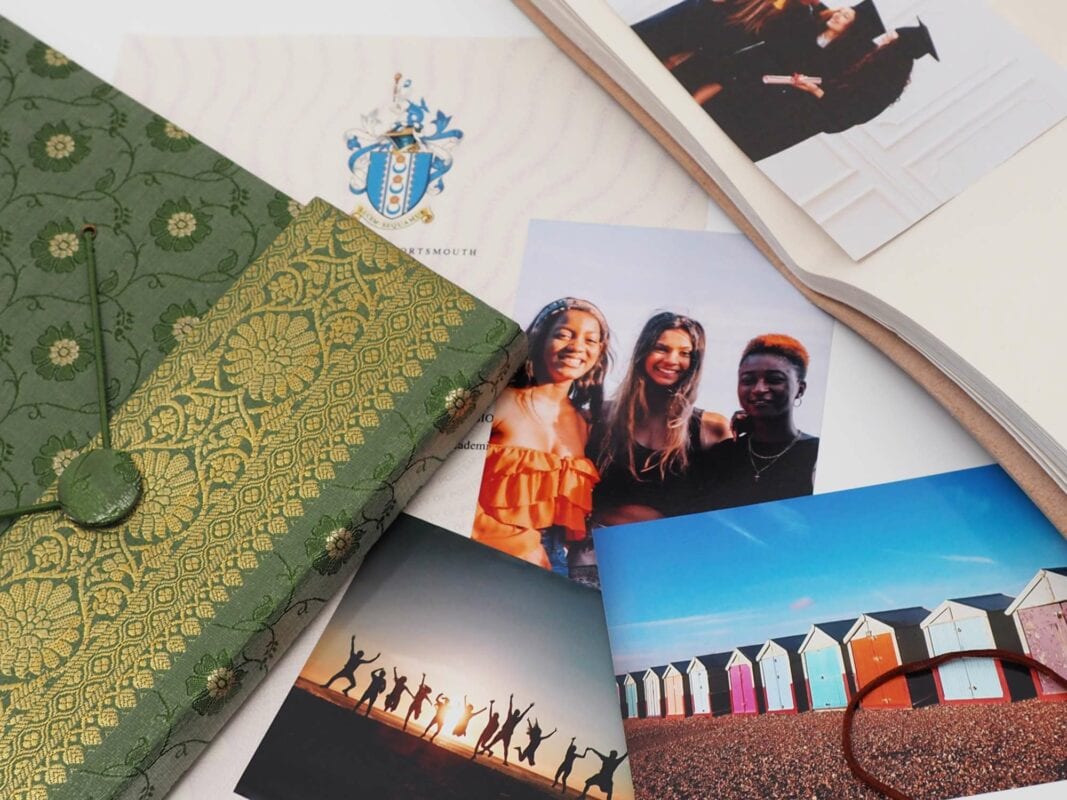 A green floral-patterned notebook, an open book, and several photographs, including three people smiling, a group jumping on a beach, and colorful beach huts featured in the Fair Trade Father's Day Gift Guide.