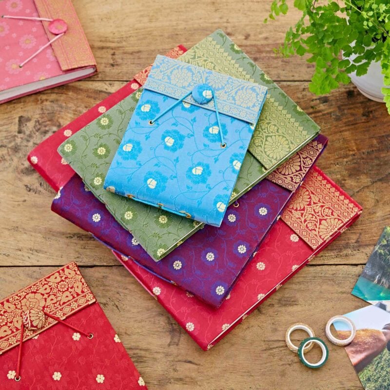 Colorful notebooks with intricate patterns reminiscent of sari fabric and a button clasp lay stacked on a wooden table. A small plant and decorative rings are placed nearby, creating an inviting scene that could easily frame a Small Sari Fabric Photo Album (Copy).