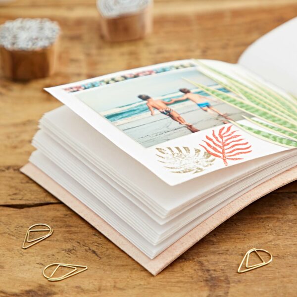 Open Indra Chakra Circle Embossed Leather Album with Semi-Precious Stones, displaying a photo of two children holding hands at the beach. The page is adorned with pressed leaves and decorative stickers, placed on a wooden table with geometric paper clips nearby. The Indra Chakra embellishment adds a touch of mysticism to the sweet memory.