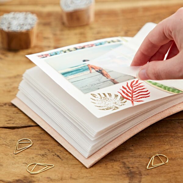 A hand places a photo of a person on a beach into the Indra Chakra Circle Embossed Leather Album with Semi-Precious Stones, which features decorative leaves. The album rests on a wooden surface, accompanied by three metal paper clips adorned with intricate designs inspired by Indra Chakra motifs.