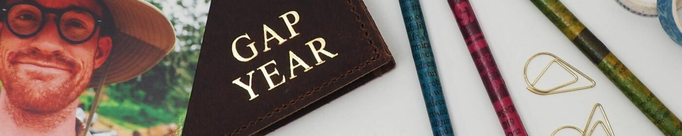Close-up of various items: a photo of a smiling person wearing glasses and a hat, a leather wallet labeled "GAP YEAR," colorful pencils, paper clips, and a ball of twine on a white surface. Perfect for your Fair Trade Father's Day Gift Guide.