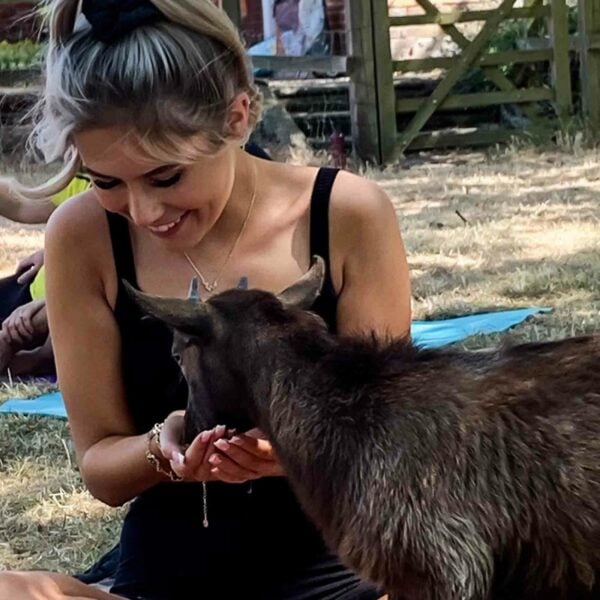 A woman, smiling and sitting outdoors on the grass, feeds a goat from her hands while enjoying a unique Goat Yoga for Two experience in the Suffolk Countryside.