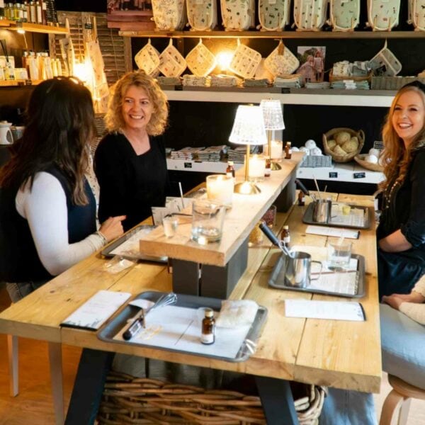 Three women sit at a wooden table in a cozy, well-lit store, engaged in conversation and smiling. The table holds various small items, trays with snacks, and drinks. Shelves with products are in the background, reflecting the store's warm ambiance as they share unlimited tea while enjoying the Art4Space "Learn The Art of Kintsugi with Unlimited Tea and Snacks" experience.