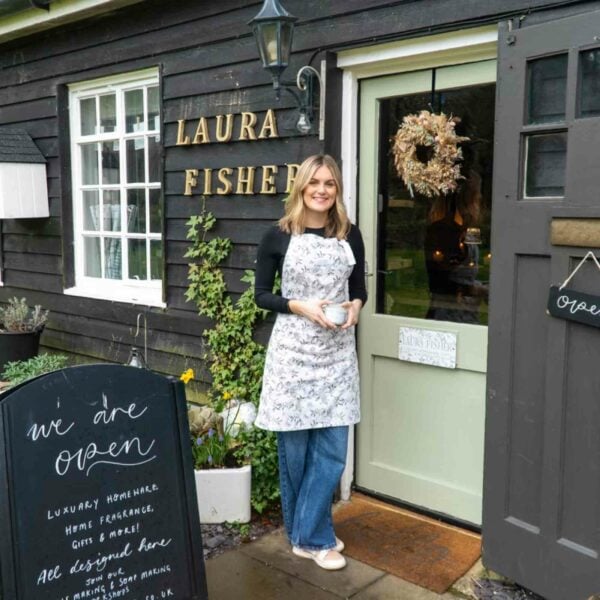 A woman in an apron stands smiling at the entrance of a shop named "Laura Fisher."