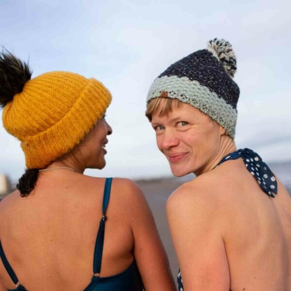 Two women wearing knitted hats look back over their shoulders while standing on a beach, as the dark sky above hints at a perfect night for enjoying the Dark Sky Wales Stargazing for Two experience.