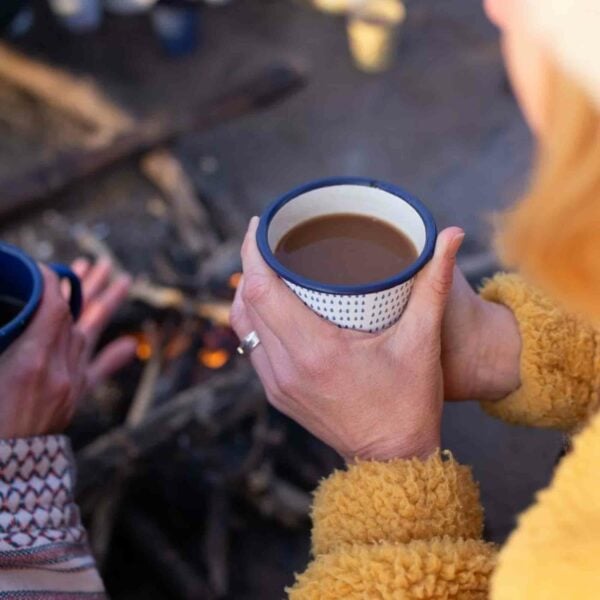 Two people holding mugs of hot beverages near a campfire, silhouetted under a dark sky. One person wears a yellow fuzzy jacket and has a ring on their left hand. Perfect for enjoying the tranquility of the night, this serene moment captures the magic of the **Dark Sky Wales Stargazing for Two** experience.