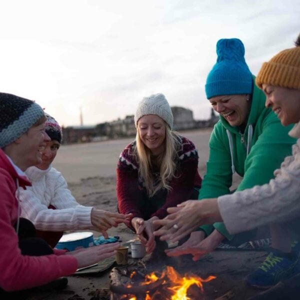 Under the vast expanse provided by Dark Sky Wales Stargazing for Two, five people dressed in winter clothing gather around a beach bonfire, warming their hands. Meanwhile, two friends break away momentarily to enjoy stargazing.