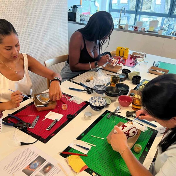 Three women are crafting at a table, surrounded by various tools and materials as they work on small projects. The backdrop reveals an urban city view through a window, highlighting their creative oasis. One of the women fondly recalls their recent adventure on the River Stour Wild Beaver Canoe Tour and Talk.