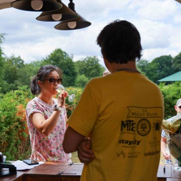 A person in sunglasses sips from a glass, possibly enjoying the Hiver's London Beekeeping and Beer Tasting Experience for Two, while another person in a yellow shirt stands nearby. They are outdoors under hanging lights with trees and greenery in the background.