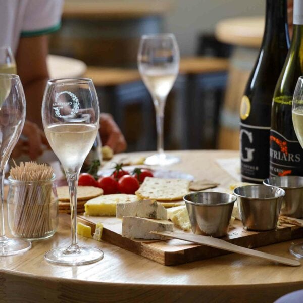 A table adorned with wine glasses, bottles of wine, cheese, crackers, and tomatoes sits ready for a delightful evening. Two empty glasses are in the foreground while a person relaxes in the background. Amidst this elegant scene reminiscent of London's charm, one might almost forget the excitement of Hiver's London Beekeeping and Beer Tasting Experience for Two awaiting them.