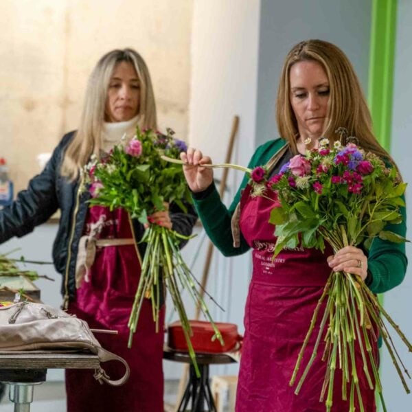 Two women, immersed in the beauty of the Suffolk Countryside, are practicing Goat Yoga for Two while arranging colorful flower bouquets in a workshop setting.