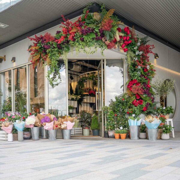 A flower shop entrance adorned with a lush garland of green, red, and yellow foliage welcomes visitors. Buckets and pots of various flowers and plants are arranged along the storefront, evoking the natural charm experienced during a session of Goat Yoga for Two in the Suffolk Countryside.