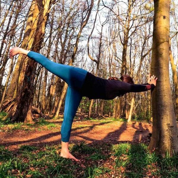 A person in blue leggings and a black top performs a yoga pose against a tree in the South Downs forest, with sunlight streaming through the trees, capturing the serenity that pairs perfectly with a "Tour the South Downs with GPS Guided Full Day Electric Mountain Bike Hire for Two.