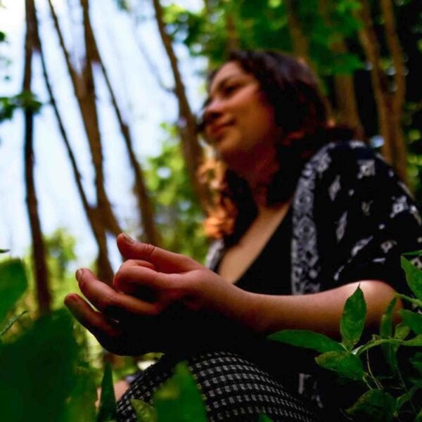 A person sits amid lush green plants in a forest, partially visible, as sunlight filters through the trees. The focus is on the foreground, with their face blurred in the background. Immerse yourself in this serene scene by opting for a GPS Guided Full Day Electric Mountain Bike Hire for Two in the South Downs, ensuring an unforgettable adventure.