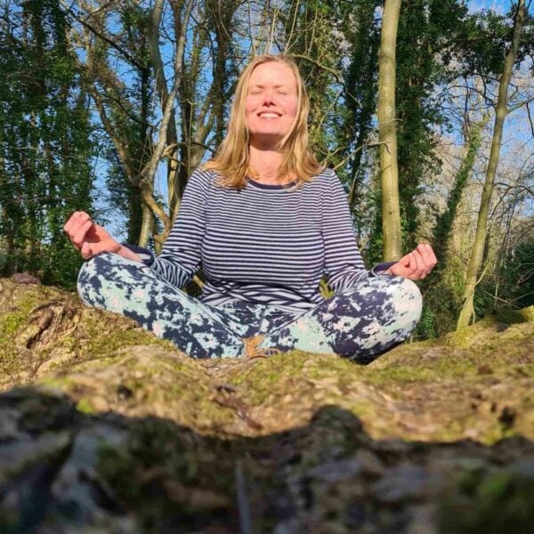 A person sits cross-legged on a rock in the South Downs, wearing a striped top and patterned leggings, eyes closed, smiling, and appearing to meditate. Nearby is the "Tour the South Downs with GPS Guided Full Day Electric Mountain Bike Hire for Two" rental spot.